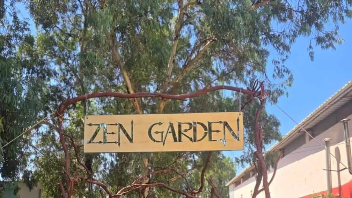 הכניסה לזן גרדן: שלט עץ ועליו הכיתוב ZEN GARDEN, תלוי על קשת ענפים מעל שביל שמוביל פנימה בין עצי אקליפטוס